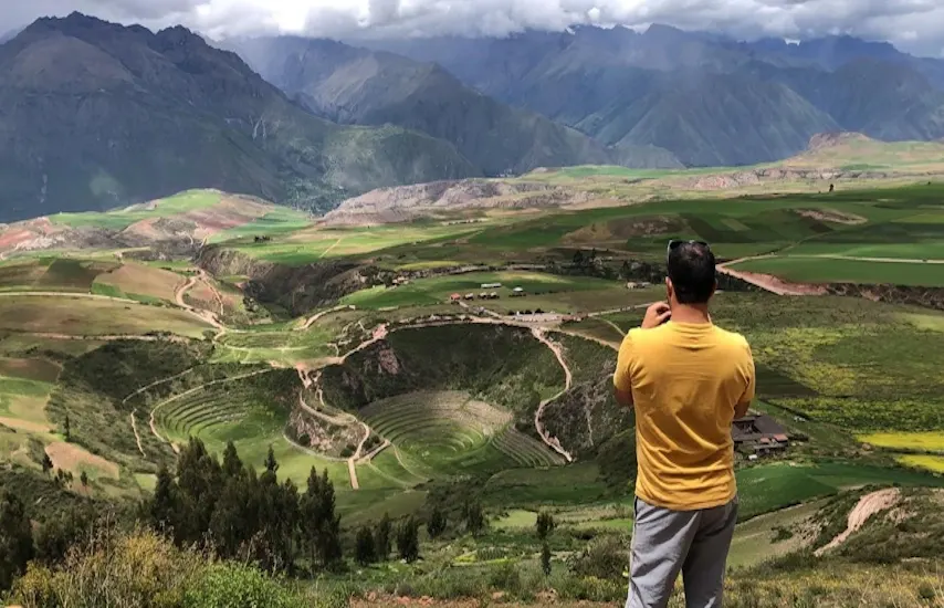 A tourist in a yellow shirt stands on a viewpoint, photographing the stunning Moray Inca agricultural terraces in the Sacred Valley of the Peruvian Andes, with layered green circular ruins surrounded by patchwork fields and snow-capped mountains under a cloudy sky.