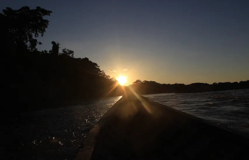 Golden sunset in the Peruvian Amazon: the sun dips behind tree silhouettes, casting its light across calm river waters as a canoe glides toward the horizon