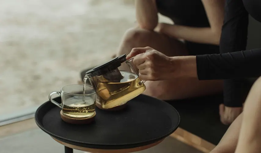 A person in black activewear pours golden herbal tea from a clear glass teapot into a matching glass mug on a black tray. The tray sits on a small round table near a window with a blurred outdoor view. The scene evokes a calm, mindful moment of relaxation.