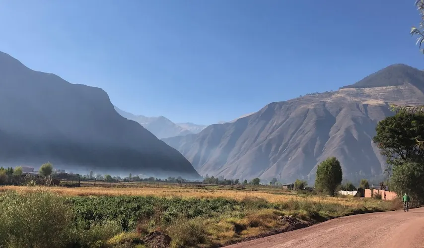 A cyclist rides along a dirt path through the Sacred Valley of the Incas, surrounded by lush green and golden fields beneath towering Andean mountains under a clear blue sky.