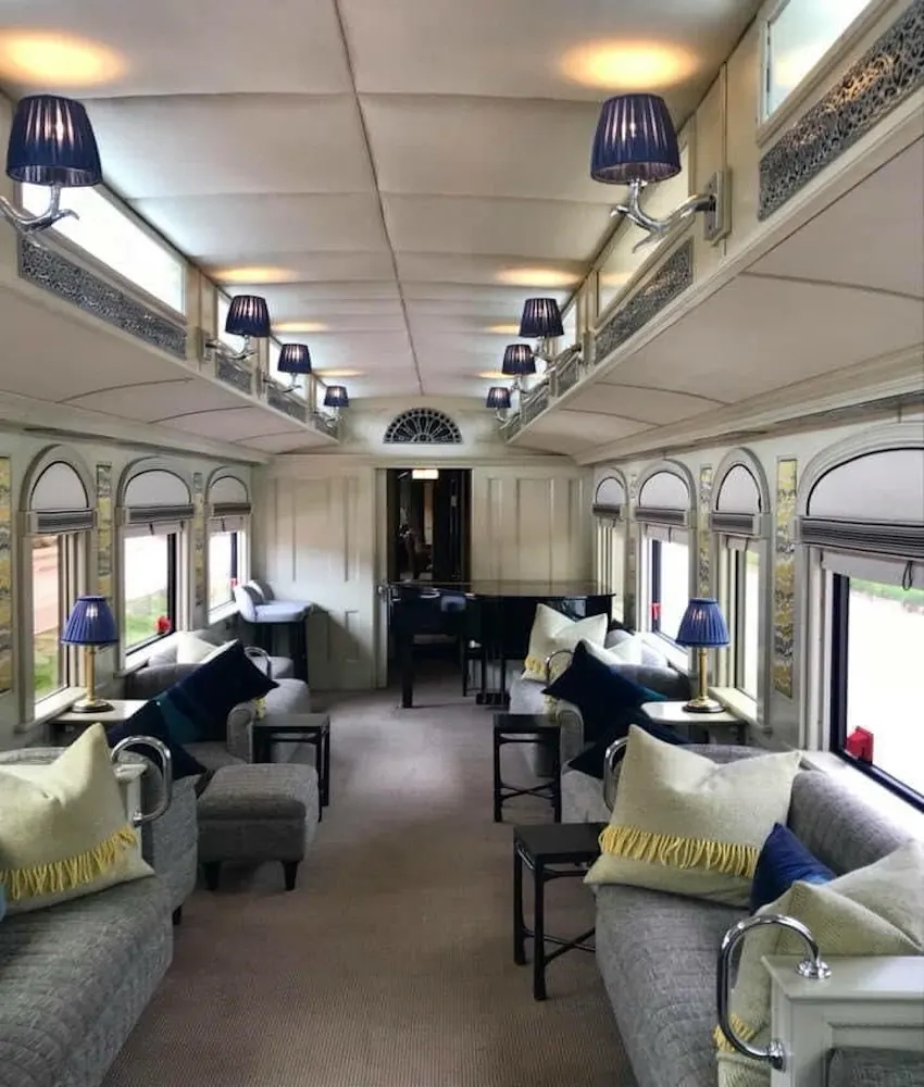 Elegant train carriage interior with plush gray sofas adorned with yellow-fringed and navy pillows, ornate wall lamps with navy shades, intricate gold-trimmed arches, and large arched windows revealing passing scenery. A grand piano sits at the far end under warm lighting.