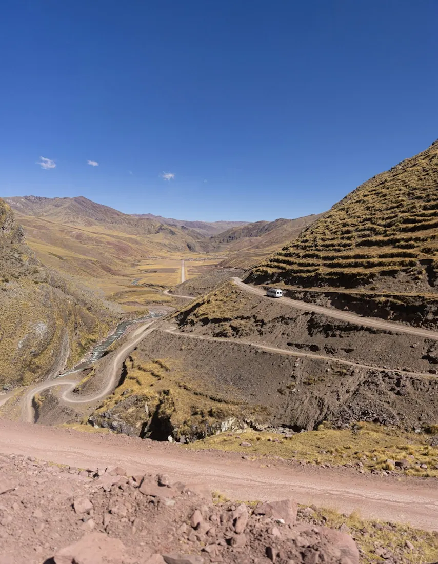 A white van travels along a winding road carved into steep slopes with golden-brown grass terraces, descending toward a narrow valley with a thin river below. Andean mountain ranges stretch out beneath a deep blue sky.