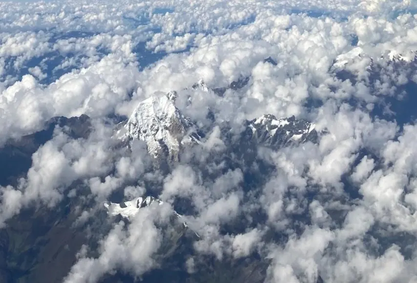 Aerial view of rugged, snow-capped Andean peaks piercing through a sea of fluffy white clouds, with distant mountain ranges fading into the horizon beneath a radiant sky.