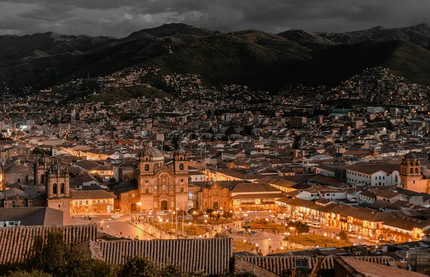 Night photograph of the city of Cusco, Peru. In the foreground, the Basilica Cathedral of the Virgin of the Assumption and the Company of Jesus in the Plaza de Armas, lit with warm light. Surrounding it, colonial buildings and cobblestone streets. In the background, Andean hills and mountains under a cloudy sky